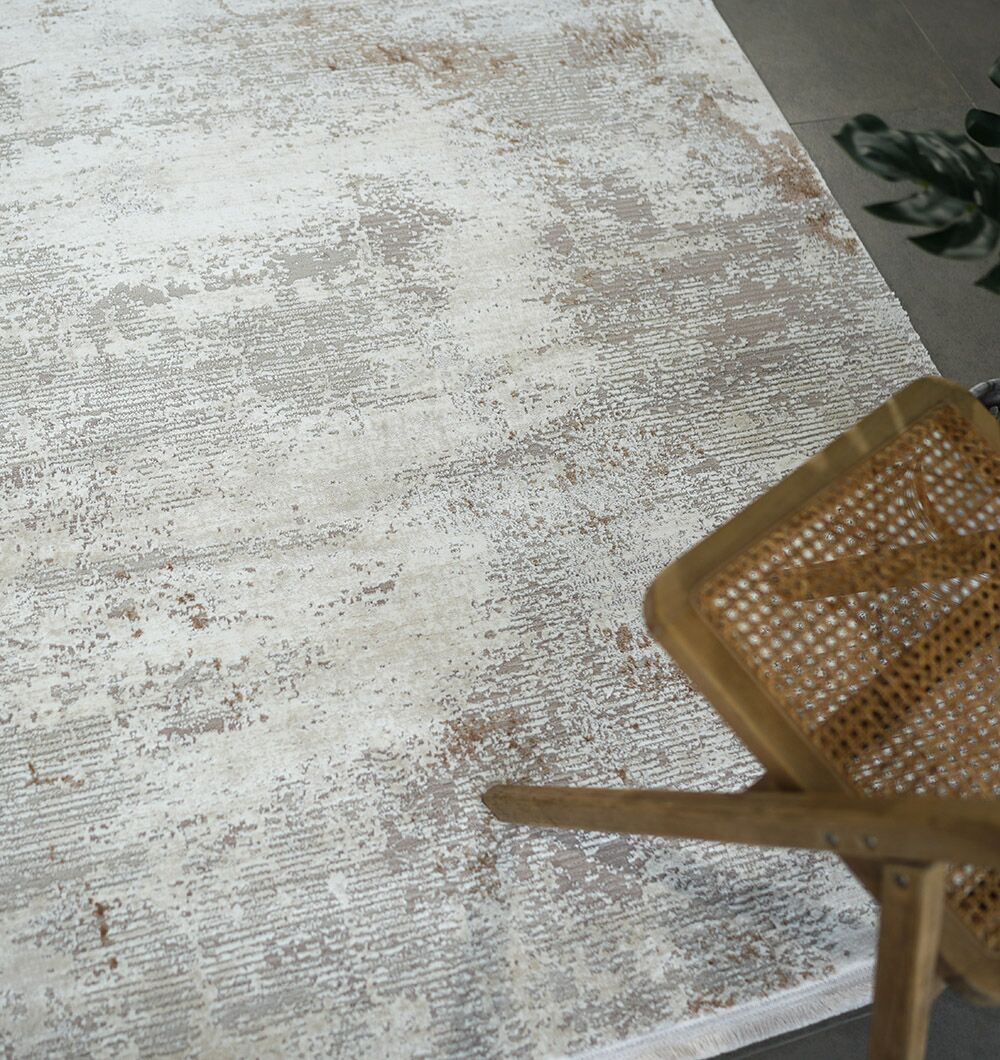 A woven chair with a cane seat sits beside the RAMSHA HOME Cream Gold Machine Made Turkish Rug - MR172 FLORENCE, showcasing subtle beige and brown patterns. A green plant appears in the upper right corner.