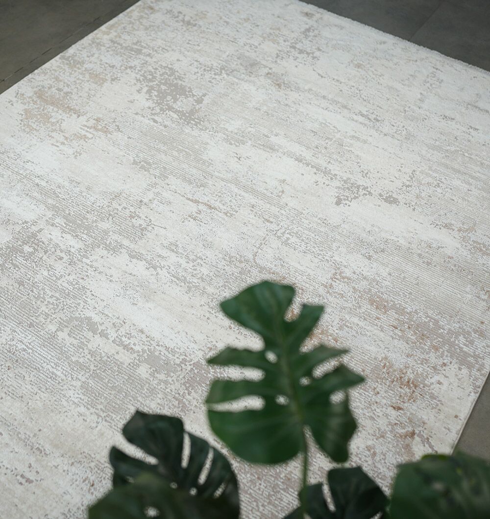 A RAMSHA HOME Cream Gold Machine Made Turkish Rug (MR172 FLORENCE) is placed on a gray floor, with green split-leaf monstera plant leaves partially visible in the foreground.