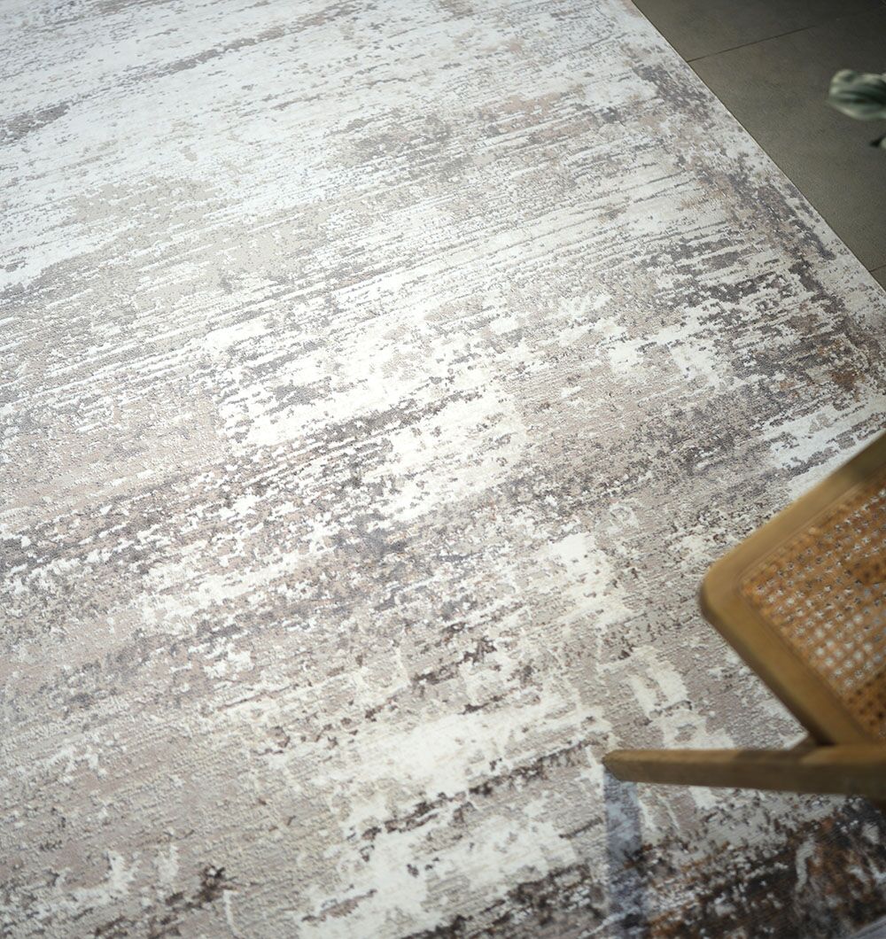A close-up of the RAMSHA HOME D.Vizon Gold Machine Made Turkish Area Rug - MR267 FLORENCE shows its modern distressed neutral pattern beside a wooden chair with a woven cane seat, highlighting the rug's elegant tones.