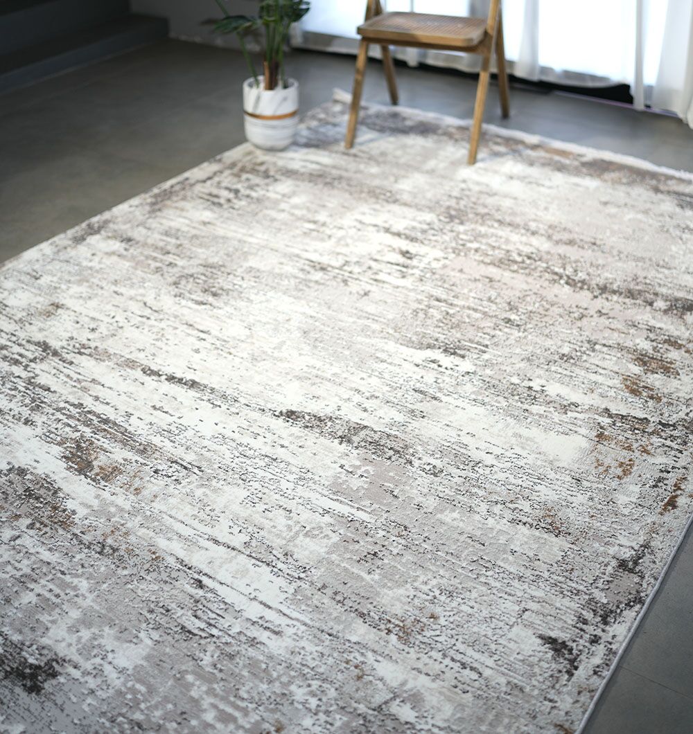 The RAMSHA HOME D.Vizon Gold Machine Made Turkish Area Rug - MR267 FLORENCE, featuring a distressed abstract pattern in grey beige, is displayed on a gray floor with a wooden chair and potted plant near a bright window.