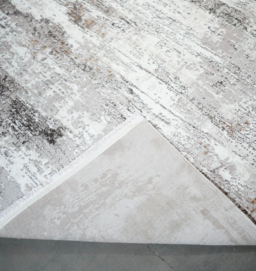 A close-up of the D.Vizon Gold Machine Made Turkish Area Rug - MR267 FLORENCE by RAMSHA HOME shows its modern neutral distressed abstract pattern in gray, white, and beige. One corner is folded over, exposing the plain underside.