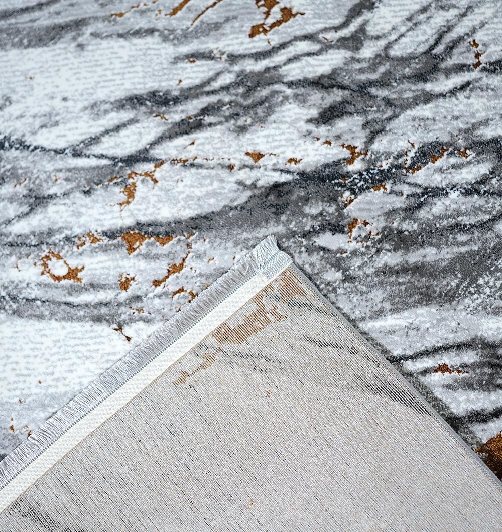 A close-up of the RAMSHA HOME Decorative Machine Made Turkish Rug - MR327 FLORENCE in grey, white, and gold shows a corner flipped over to highlight its marble-look texture and detailed underside.