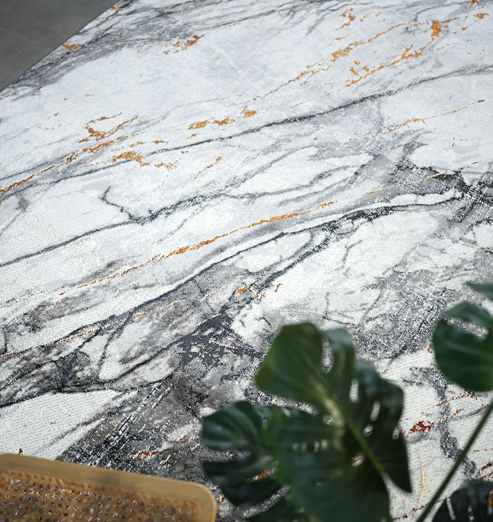 A close-up of the RAMSHA HOME Decorative Machine Made Turkish Rug - MR327 FLORENCE features white, gray, and gold marble veining, with a green leafy plant in the lower right and a woven chair edge in the lower left.