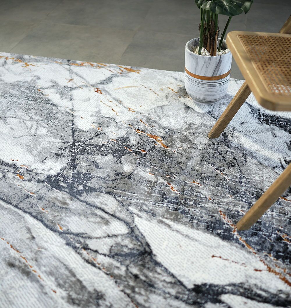 The RAMSHA HOME Decorative Machine Made Turkish Rug - MR327 FLORENCE, featuring a marbled gray, white, and subtle orange pattern, lies on a tiled floor beside a potted plant and the corner of a woven wooden chair.