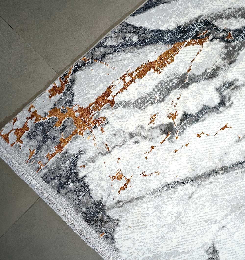 A close-up of the RAMSHA HOME Decorative Machine Made Turkish Rug - MR327 FLORENCE, featuring a modern marble-like pattern in white, gray, black, and gold tones. This rectangular rug has fringed edges on one side and is displayed on a tiled floor.