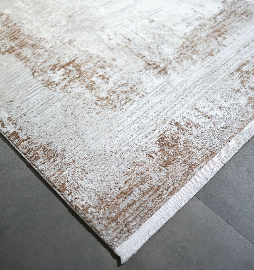 A close-up of the RAMSHA HOME MR359 FLORENCE, a machine made Turkish rug in cream and gold, featuring a textured abstract pattern, displayed on a smooth gray tile floor.