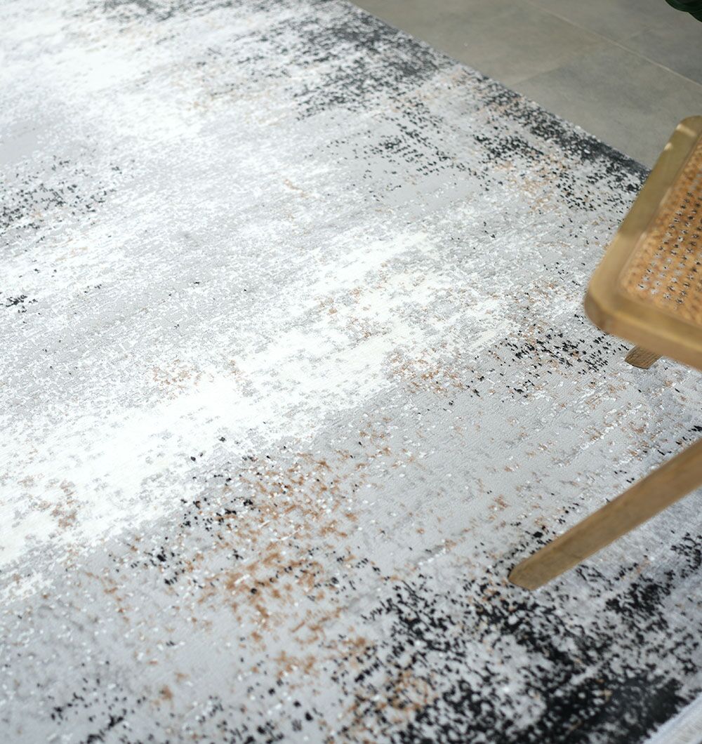 A close-up of the RAMSHA HOME MR529 FLORENCE Machine Made Turkish Area Rug shows its mottled white, gray, black, and beige pattern. A wooden chair with a woven seat in the corner highlights the rug's contemporary neutral style.