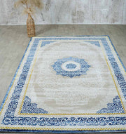 The RAMSHA HOME Machine Made Turkish Area Rug - N370A MADRID, featuring a beige background with ornate blue and gold borders and a blue medallion center, is placed on a light wood floor. A tall vase with pampas grass sits in the back left corner by a textured wall.