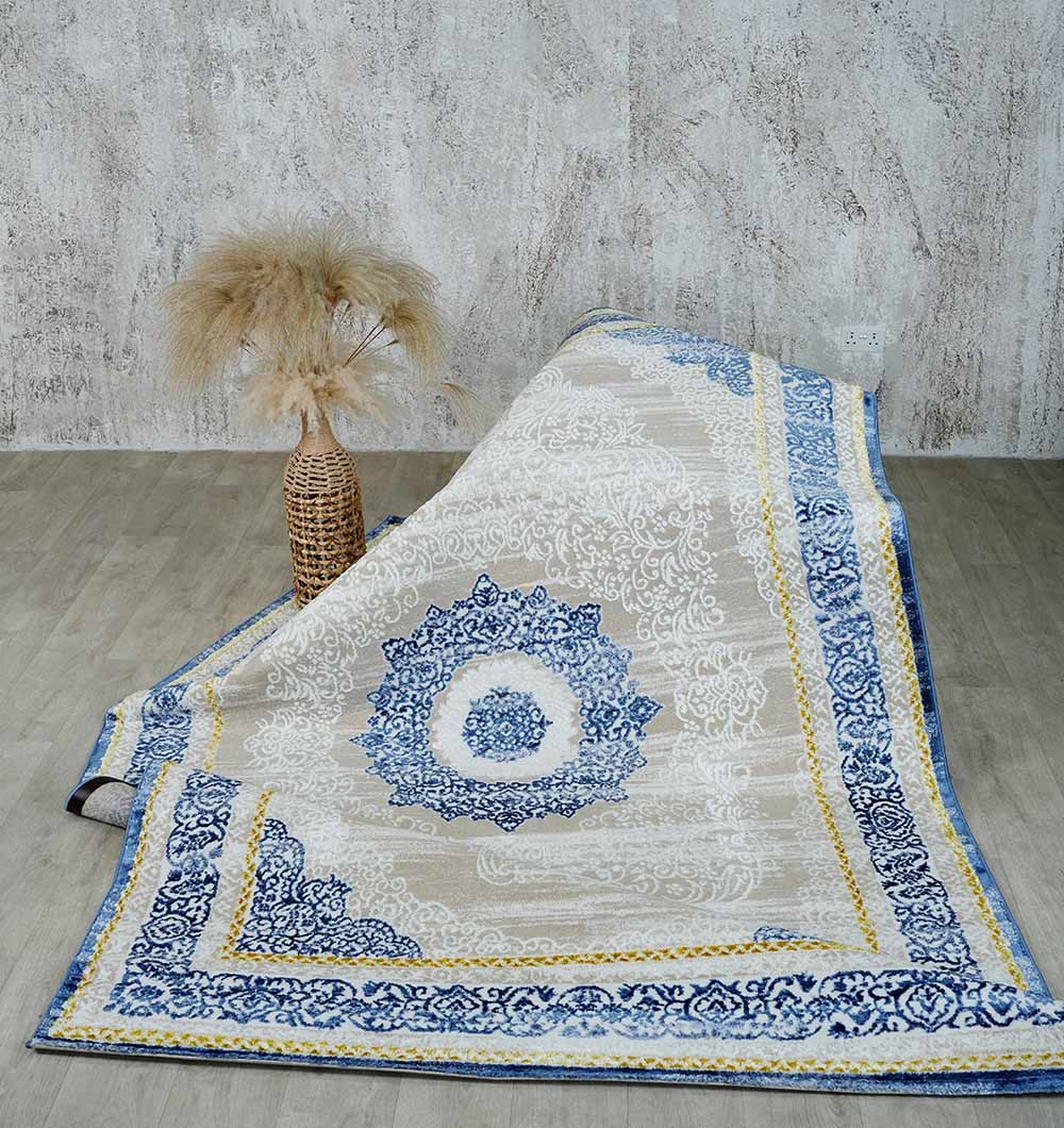 The RAMSHA HOME Machine Made Turkish Area Rug - N370A MADRID in blue, gold, and white with ornate patterns is displayed beside a tall vase of dried pampas grass, set against a textured gray wall and light wood flooring.