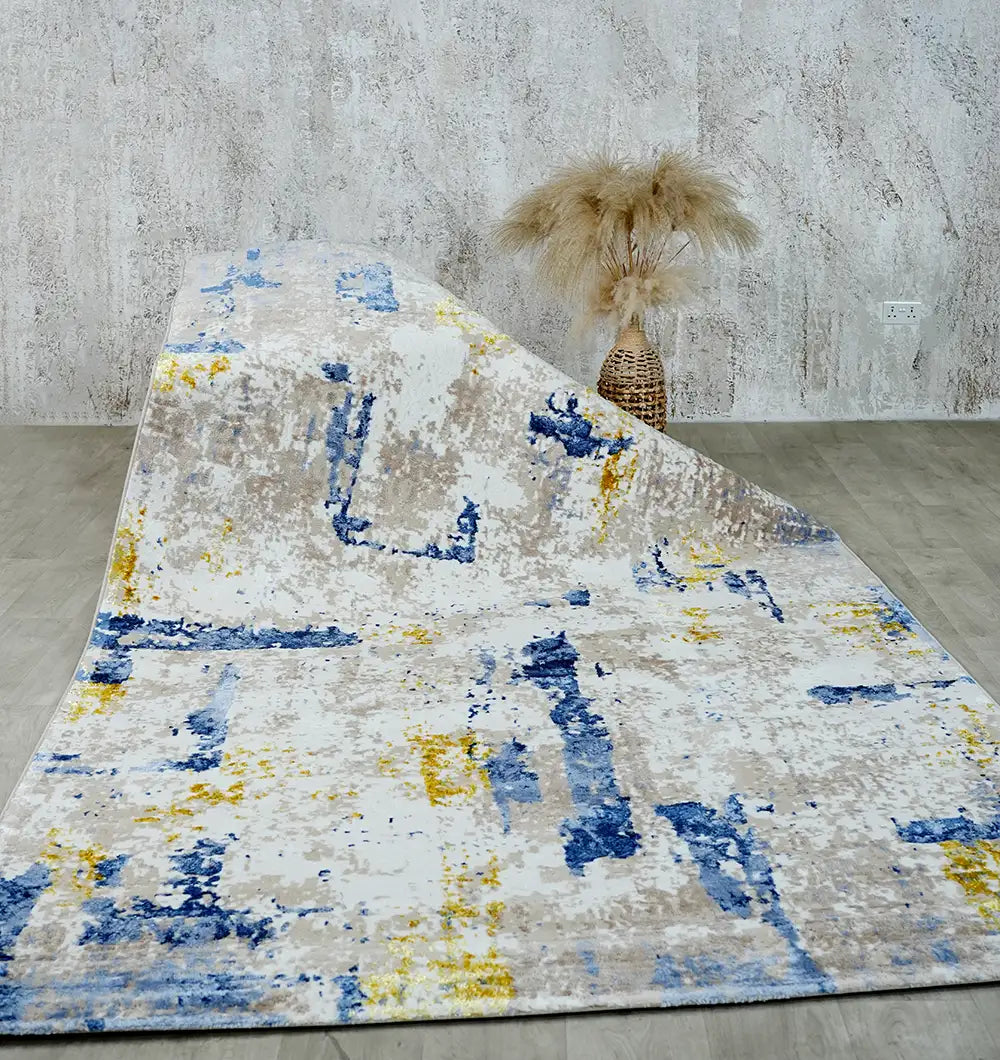The RAMSHA HOME Machine Made Turkish Area Rug - N371A MADRID, featuring abstract blue, yellow, and gray patterns, is partially draped across a light wood floor. Pampas grass in a woven vase accents the textured gray wall in the background.