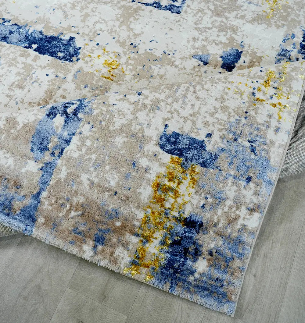 The RAMSHA HOME Machine Made Turkish Area Rug - N371A MADRID features a modern abstract design in blue, yellow, and beige on a light background, shown here partially folded at one corner on a light wood floor.