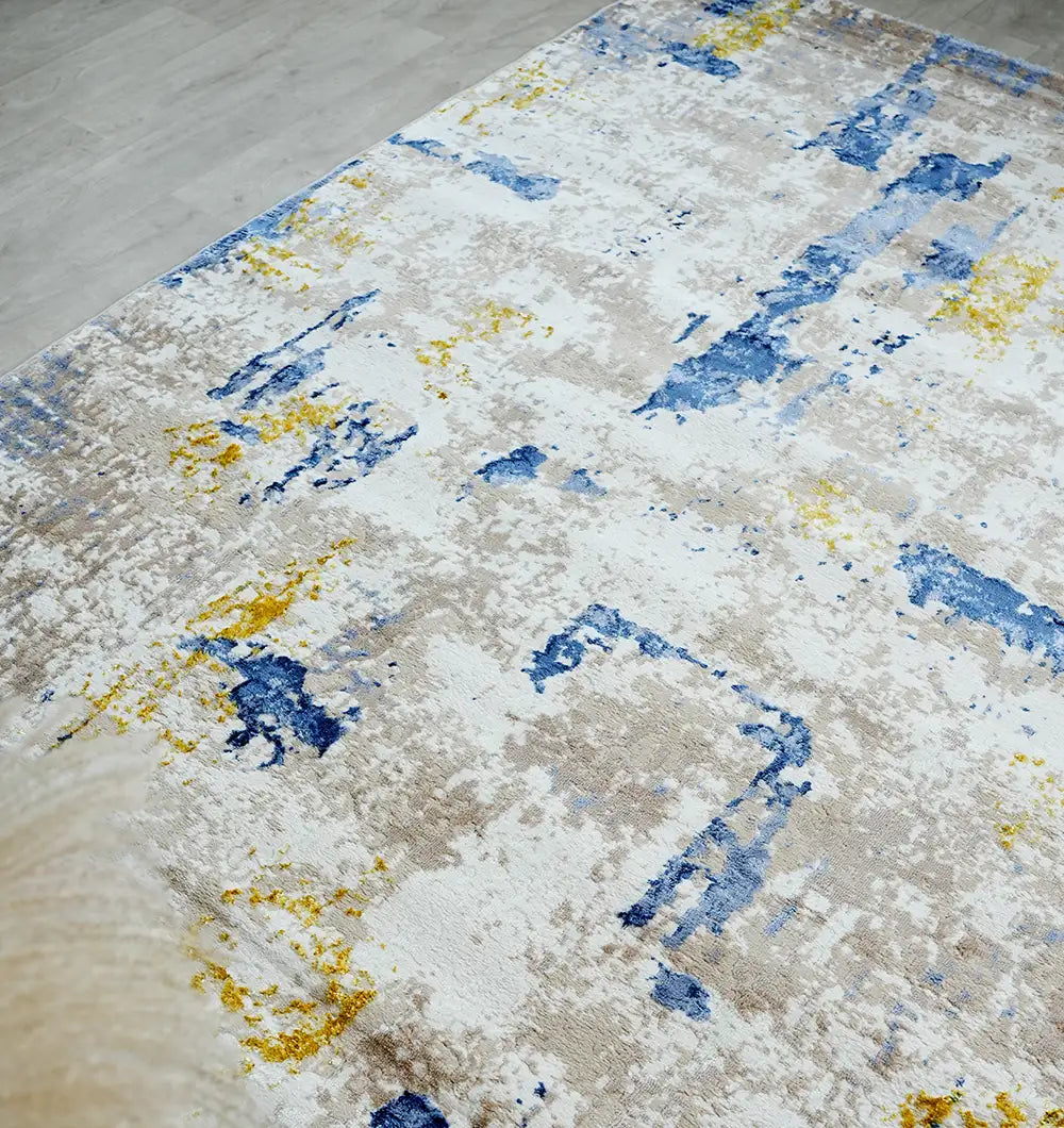 A close-up of the RAMSHA HOME Machine Made Turkish Area Rug - N371A MADRID, featuring an abstract pattern in blue, yellow, beige, and white on a light-colored floor.
