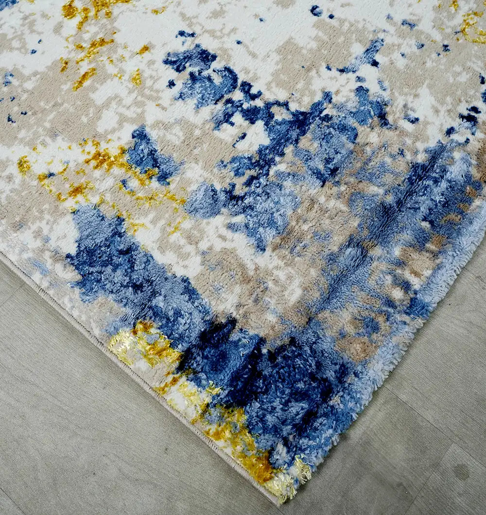 The RAMSHA HOME Machine Made Turkish Area Rug - N371A MADRID features an abstract blue, yellow, and beige pattern with plush fibers and textured patches, displayed on a light gray wooden floor for a modern industrial look.