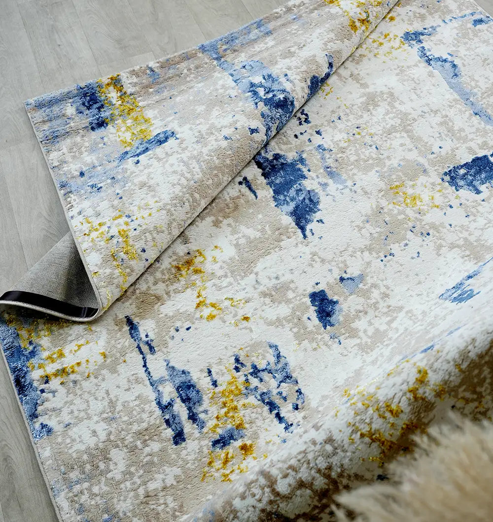 The RAMSHA HOME Machine Made Turkish Area Rug - N371A MADRID features a modern abstract pattern in blue, yellow, gray, and white with a textured look, shown partially folded on a light wood floor.