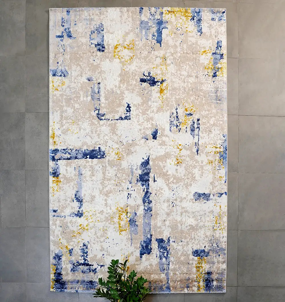 The RAMSHA HOME Machine Made Turkish Area Rug - N371A MADRID, featuring an abstract geometric pattern in blue, yellow, and beige, lies on a gray tiled floor. A green leafy plant appears partially at the bottom center of the image.
