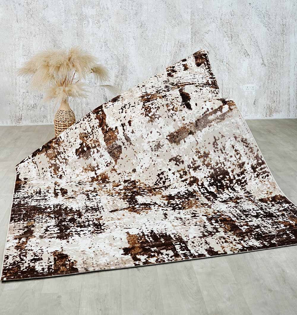 The RAMSHA HOME Beige & Dark Brown Turkish Area Rug - N371B MADRID, featuring abstract designs, is partially folded on a light wood floor. A woven vase with pampas grass sits by a textured light gray wall in the background.