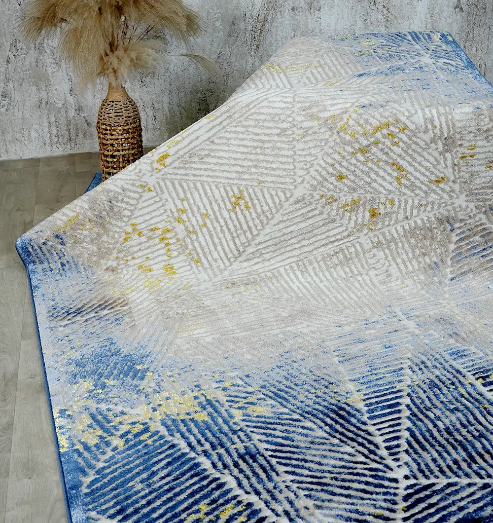 The RAMSHA HOME Abstract Geometric Machine Made Turkish Area Rug - N405A MADRID, featuring a modern leaf pattern in blue, beige, and yellow tones, lies on a wood floor beside a tall wicker vase with dried pampas grass against a gray wall.
