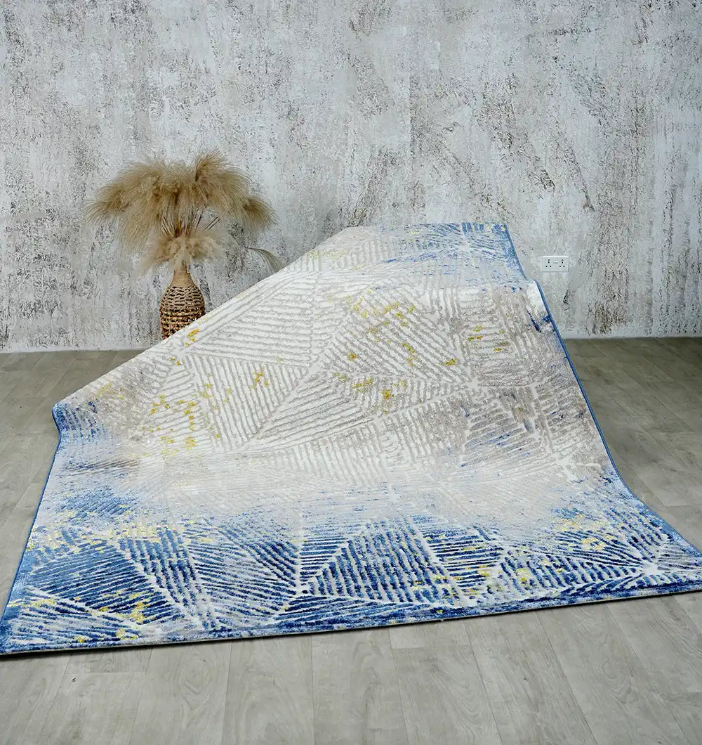 The RAMSHA HOME Abstract Geometric Machine Made Turkish Area Rug - N405A MADRID in blue and white with striking patterns is draped on a light wooden floor, while a vase of dried pampas grass stands before a textured gray wall.