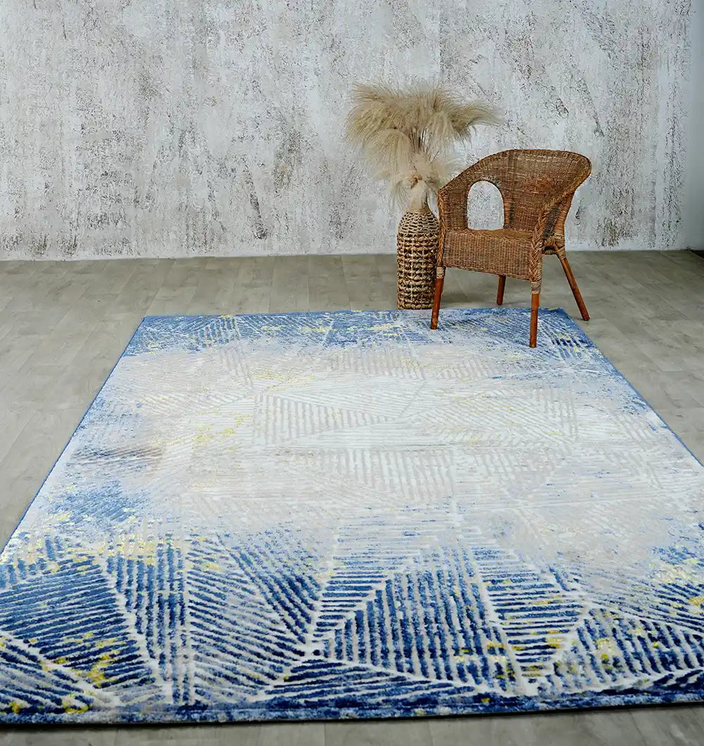 A modern room showcases the RAMSHA HOME Abstract Geometric Machine Made Turkish Area Rug - N405A MADRID in blue and white on a light wooden floor, paired with a wicker chair and tall pampas grass beside a textured gray wall.