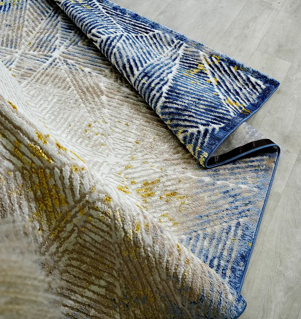 The RAMSHA HOME Abstract Geometric Machine Made Turkish Area Rug - N405A MADRID, featuring blue, white, gray, and yellow patterns, is partially folded on a light wood floor to display both its front and underside.