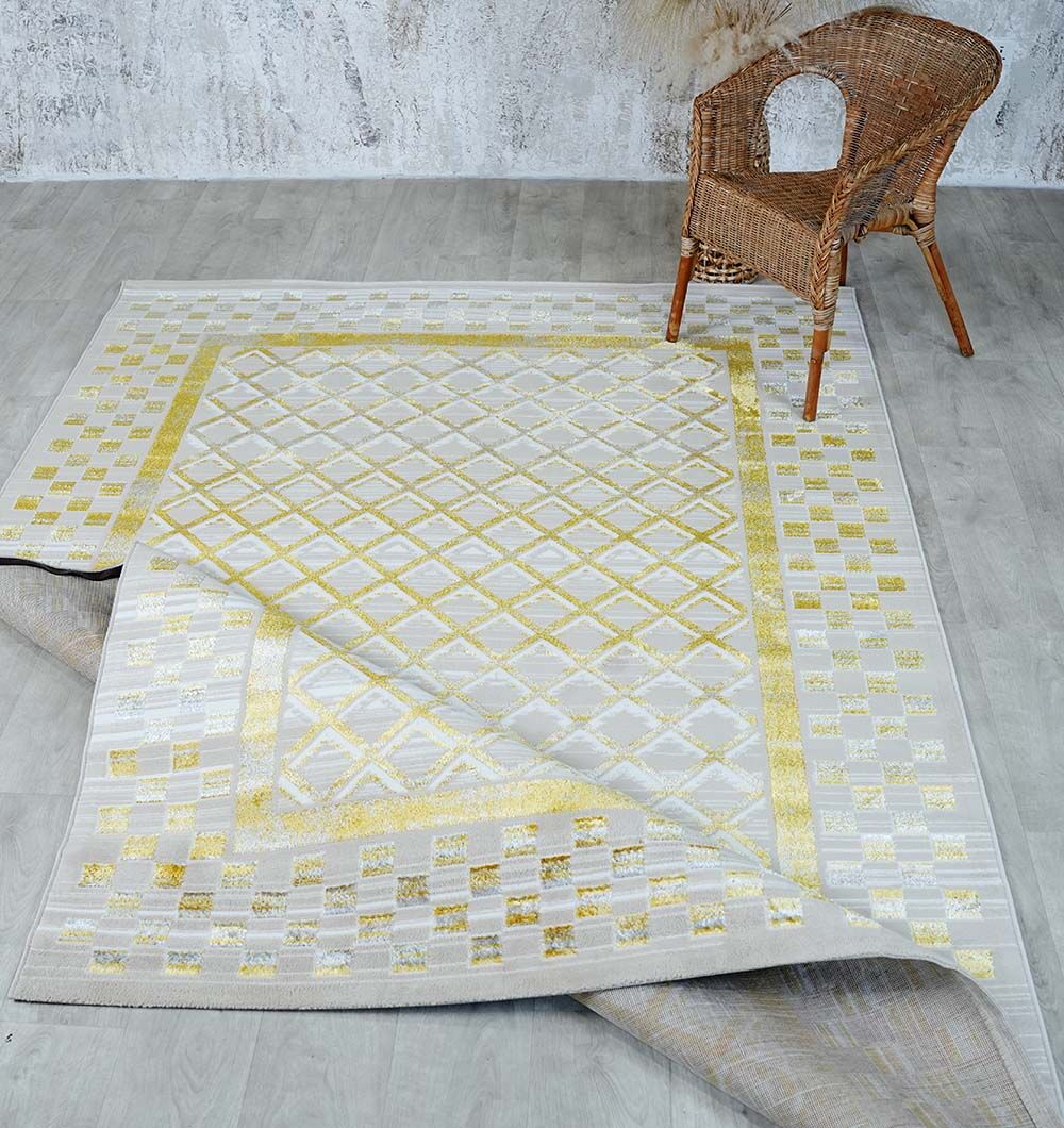A woven wicker chair sits on a light gray floor next to the RAMSHA HOME Beige & Gold Silk-Feel Turkish Rug (NK71A MADRID), which features a geometric pattern. One corner of the rug is folded over, showing its underside.