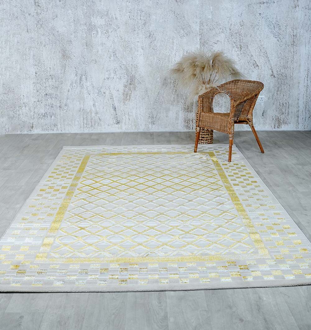 A RAMSHA HOME Beige & Gold Silk-Feel Turkish Rug (NK71A MADRID) lies on a light wood floor. In the corner, a wicker chair and a vase with dried pampas grass sit against a textured gray wall.