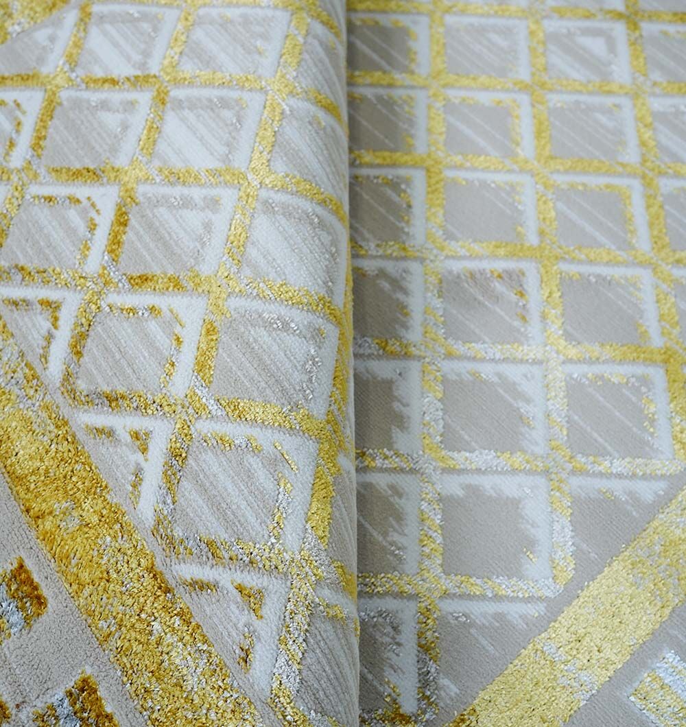 A close-up of the RAMSHA HOME Beige & Gold Silk-Feel Turkish Rug - NK71A MADRID showcases its shiny gold geometric square pattern and luxurious texture, with a gentle central fold highlighting its plush thickness.
