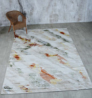 A neutral-toned room with light wood flooring showcases the RAMSHA HOME Machine Made Turkish Decor Rug - NK73A MADRID, featuring abstract orange, yellow, gray, and green patterns. A wicker chair sits in the top left beside a vase with branches.