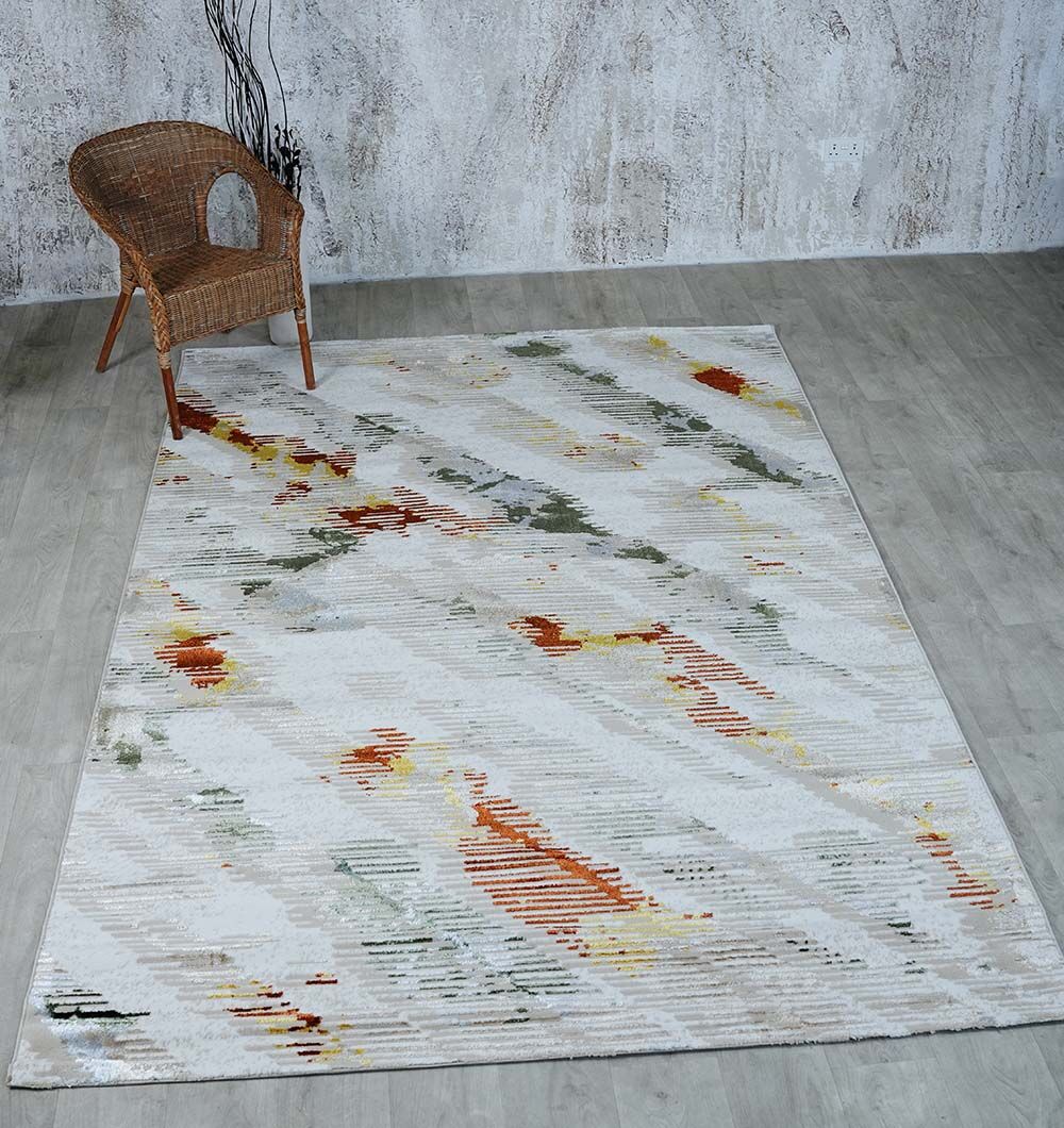 A neutral-toned room with light wood flooring showcases the RAMSHA HOME Machine Made Turkish Decor Rug - NK73A MADRID, featuring abstract orange, yellow, gray, and green patterns. A wicker chair sits in the top left beside a vase with branches.