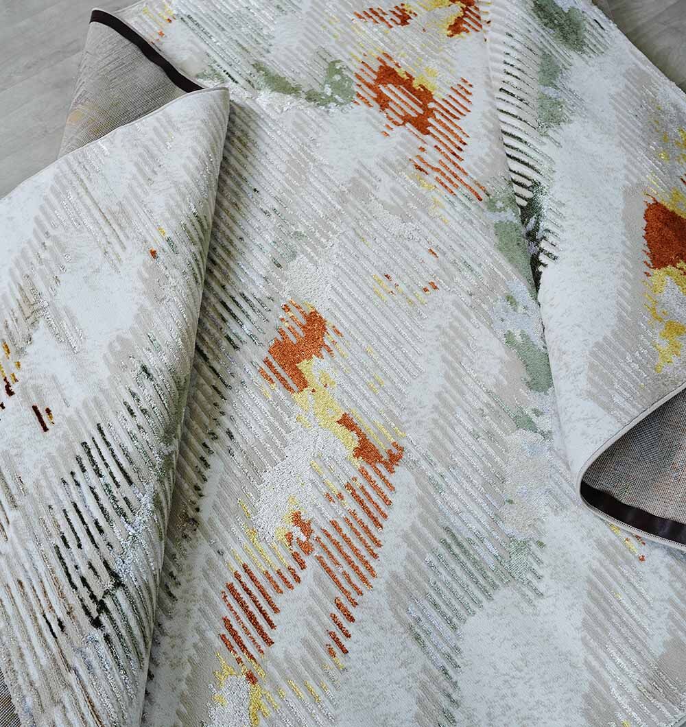 A close-up of the RAMSHA HOME Machine Made Turkish Decor Rug - NK73A MADRID, featuring abstract orange, yellow, and green patterns on a light background, partially folded to highlight its texture and underside.