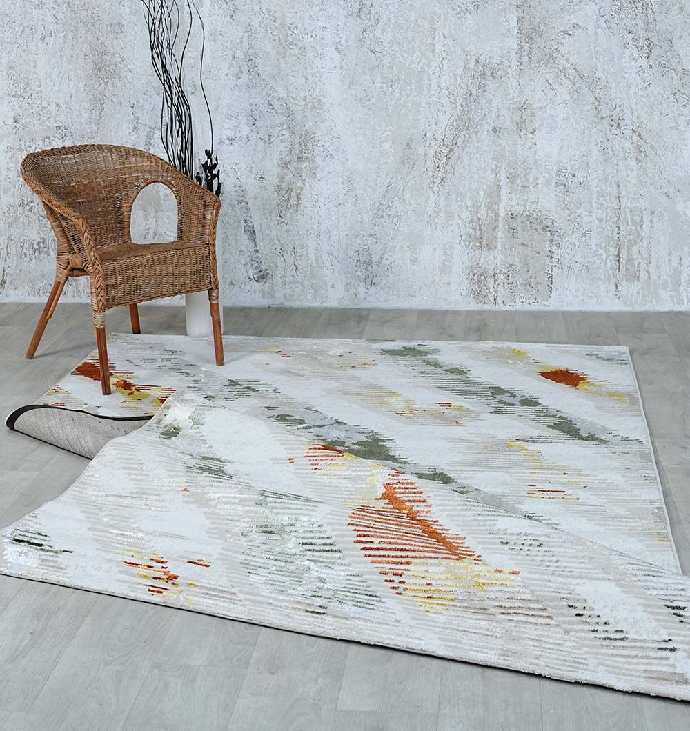 The RAMSHA HOME Machine Made Turkish Decor Rug - NK73A MADRID features light hues with abstract orange, yellow, and gray patterns. Partially folded at one corner, it sits under a wicker chair against a textured light gray wall and wood floor.