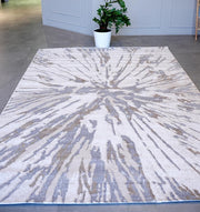The RAMSHA HOME Abstract Sunburst Machine Made Turkish Decor Rug - NK79A MADRID, featuring a light cream background with gray and beige bursts, is displayed on a tiled floor with a potted plant and light gray walls in the background.