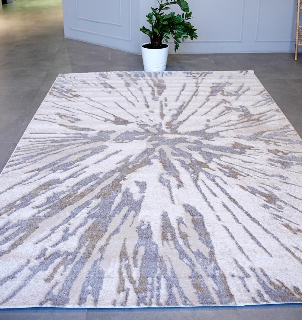 The RAMSHA HOME Abstract Sunburst Machine Made Turkish Decor Rug - NK79A MADRID, featuring a light cream background with gray and beige bursts, is displayed on a tiled floor with a potted plant and light gray walls in the background.