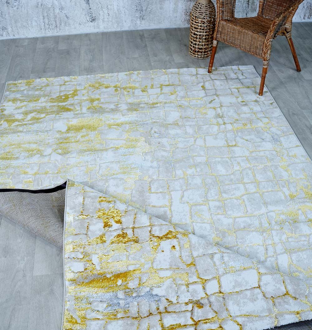The RAMSHA HOME Abstract Machine Made Ivory & Yellow Turkish Area Rug - NK81A MADRID is partially folded on a light wood floor, near a wicker chair and woven basket, set against a textured gray wall.