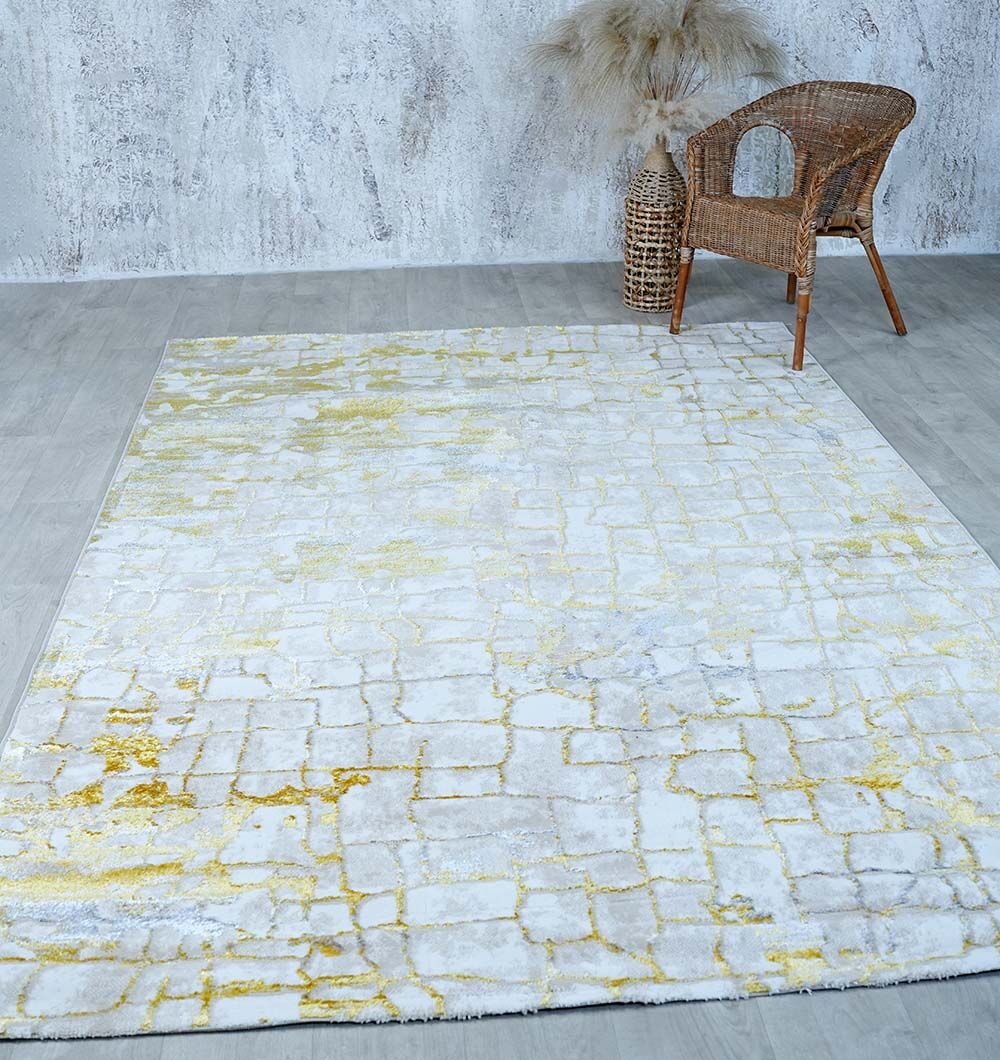A modern room with a RAMSHA HOME Abstract Machine Made Ivory & Yellow Turkish Area Rug (NK81A MADRID), featuring subtle Persian motifs, a wicker chair, pampas grass in a tall vase, textured white wall, and light wood flooring.