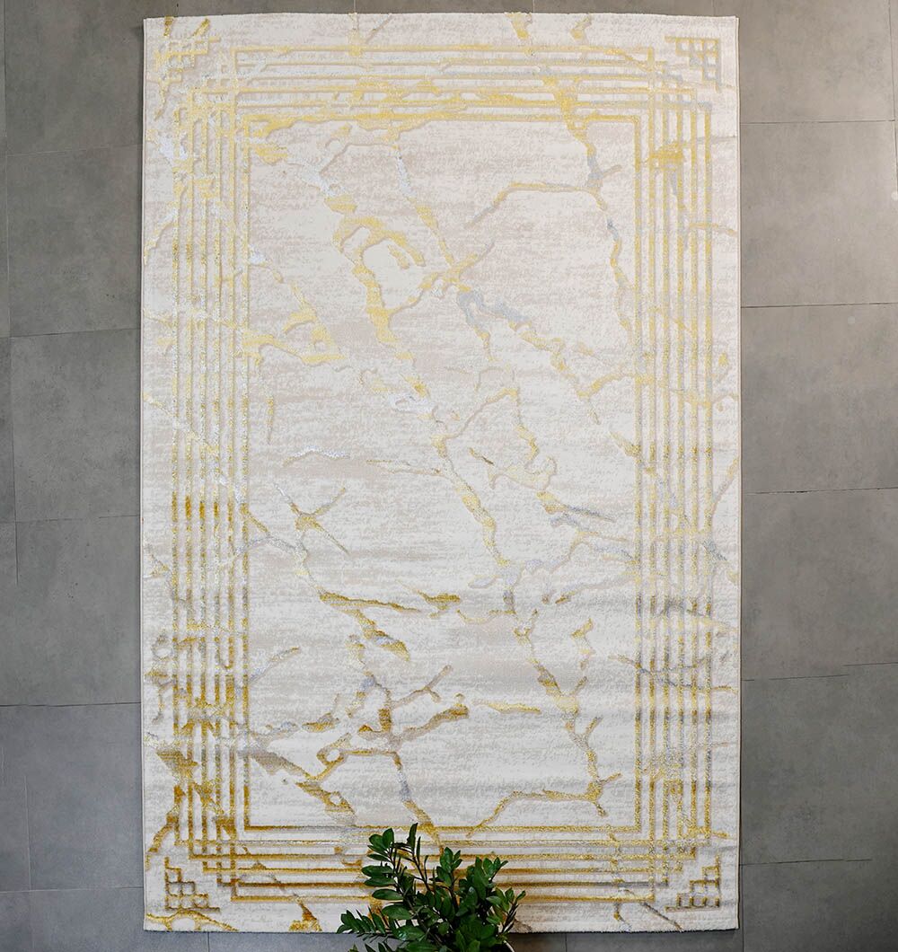 The RAMSHA HOME Ivory & Gold Machine Made Turkish Area Rug - NK82A MADRID features a modern rectangular design with a light cream base, gold marble veining, and a geometric gold border, displayed on a gray wall above a green leafy plant.
