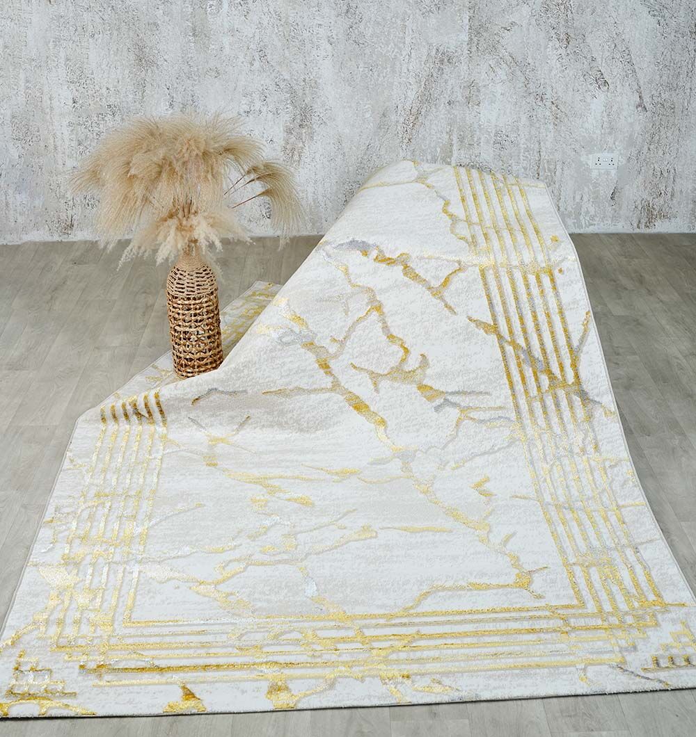 The RAMSHA HOME Ivory & Gold Machine Made Turkish Area Rug - NK82A MADRID lies on a light wood floor, paired with a tall woven vase of pampas grass, all set against a textured gray wall for an elegant look.