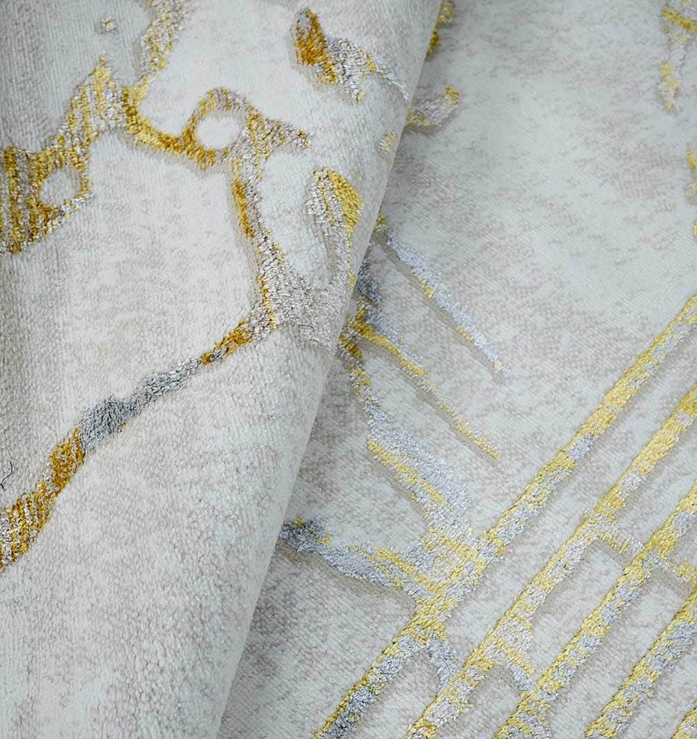 A close-up of the RAMSHA HOME Ivory & Gold Machine Made Turkish Area Rug - NK82A MADRID displays abstract gold and silver metallic patterns on light gray, with a textured surface and subtle folds highlighting its shimmer and intricate design.