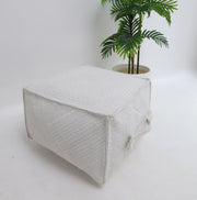 A RAMSHA HOME Bleach color Floor Square Pouf with Handle (PF 088) features a textured design and sits on a white floor next to a cream-colored pot with a leafy green plant, providing stylish extra seating ideal for modern interiors.