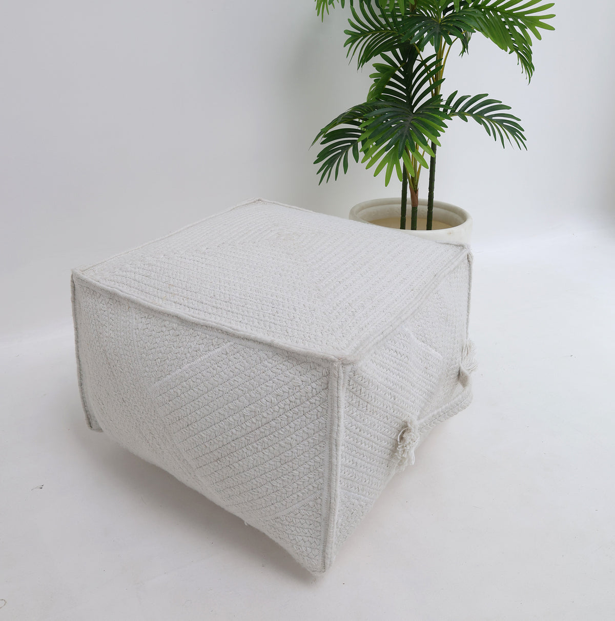 A RAMSHA HOME Bleach color Floor Square Pouf with Handle (PF 088) features a textured design and sits on a white floor next to a cream-colored pot with a leafy green plant, providing stylish extra seating ideal for modern interiors.
