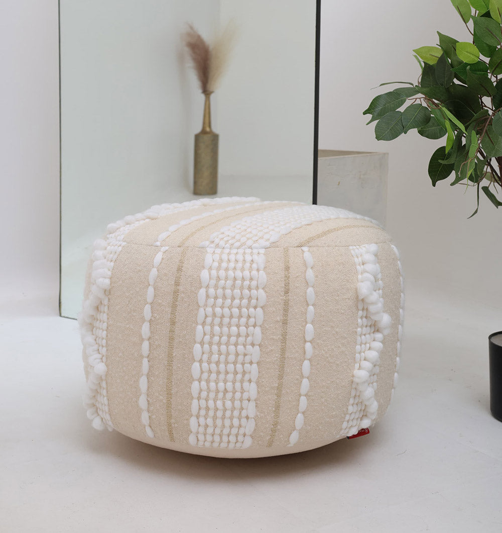 The RAMSHA HOME Boho Modern Round Pouf With Insert PF 090, cream-colored with textured white stripes and bobbles, adds extra seating on a white floor. Behind it, a mirror reflects a vase of dried grasses and a nearby green plant.