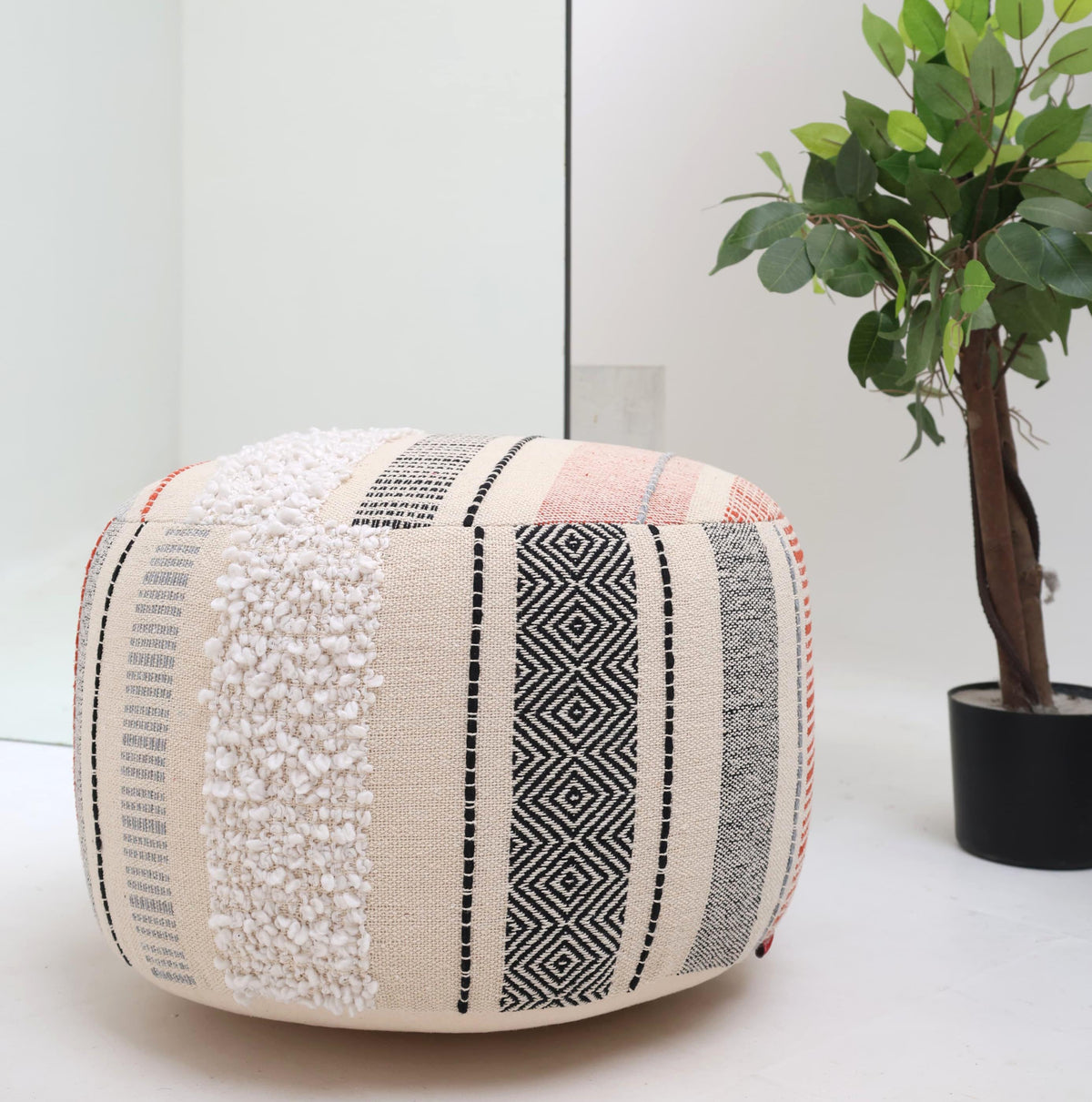 A RAMSHA HOME Boho Modern Round Pouf With Insert PF 105, featuring beige cotton fabric with black geometric and textured white patterns, sits on a white floor next to a green potted plant and in front of a large mirror, providing extra seating.
