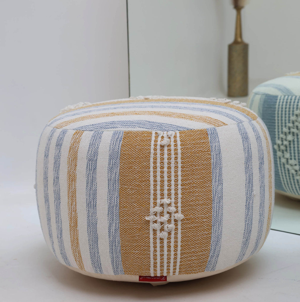 The Boho Modern Round pouf With Insert PF 109 from RAMSHA HOME, featuring yellow and blue vertical stripes with textured white details, serves as a charming decorative accent and provides extra seating when placed in front of a mirror on a white surface.