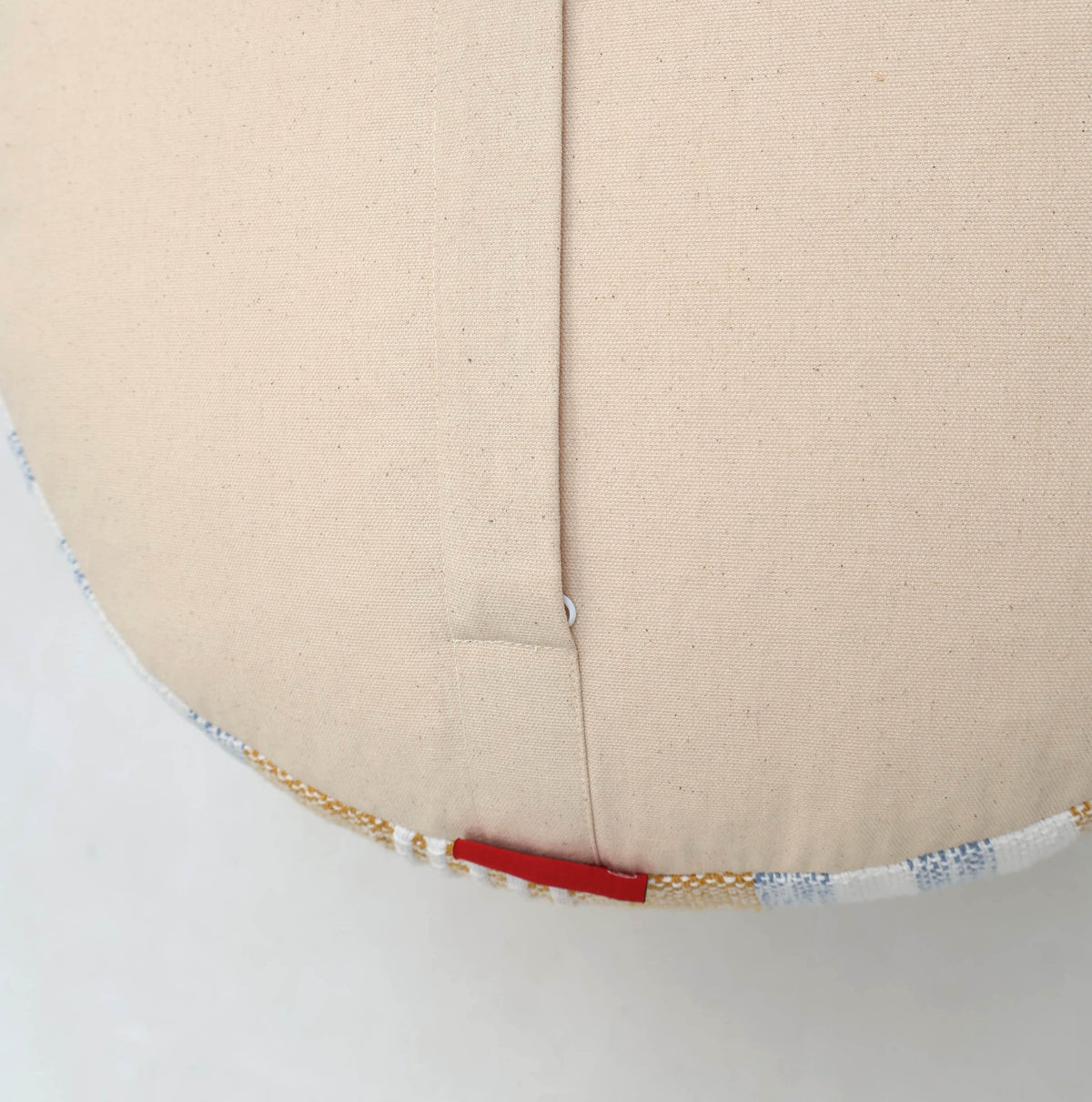 A close-up of the Boho Modern Round pouf With Insert PF 109 by RAMSHA HOME showcases its beige fabric design featuring a central seam, a small gold chain accent, and a short red bar stitched near the edge—making it an elegant decorative accent piece for any room.