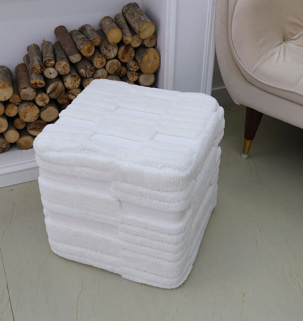 A stack of white, textured towels sits on the floor beside the RAMSHA HOME Handmade Pouf with Insert White Color PF-152, a light armchair, and a pile of firewood stacked against the wall.