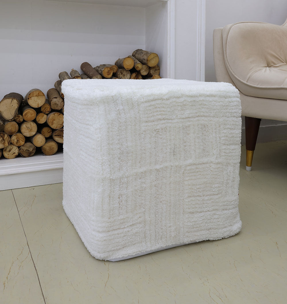 The RAMSHA HOME Handmade Pouf with Insert White Color PF-154, featuring soft textured fabric and a thermocol cube insert, adds a premium touch to your space when placed on a light floor near an armchair and firewood against a white wall.
