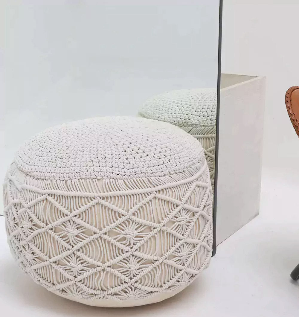 The RAMSHA HOME Boho Macrame Pouf White Round PF-155, featuring a textured diamond pattern, sits before a mirror and reflects its stylish design. A brown chair is partly visible on the right against the minimal white background.