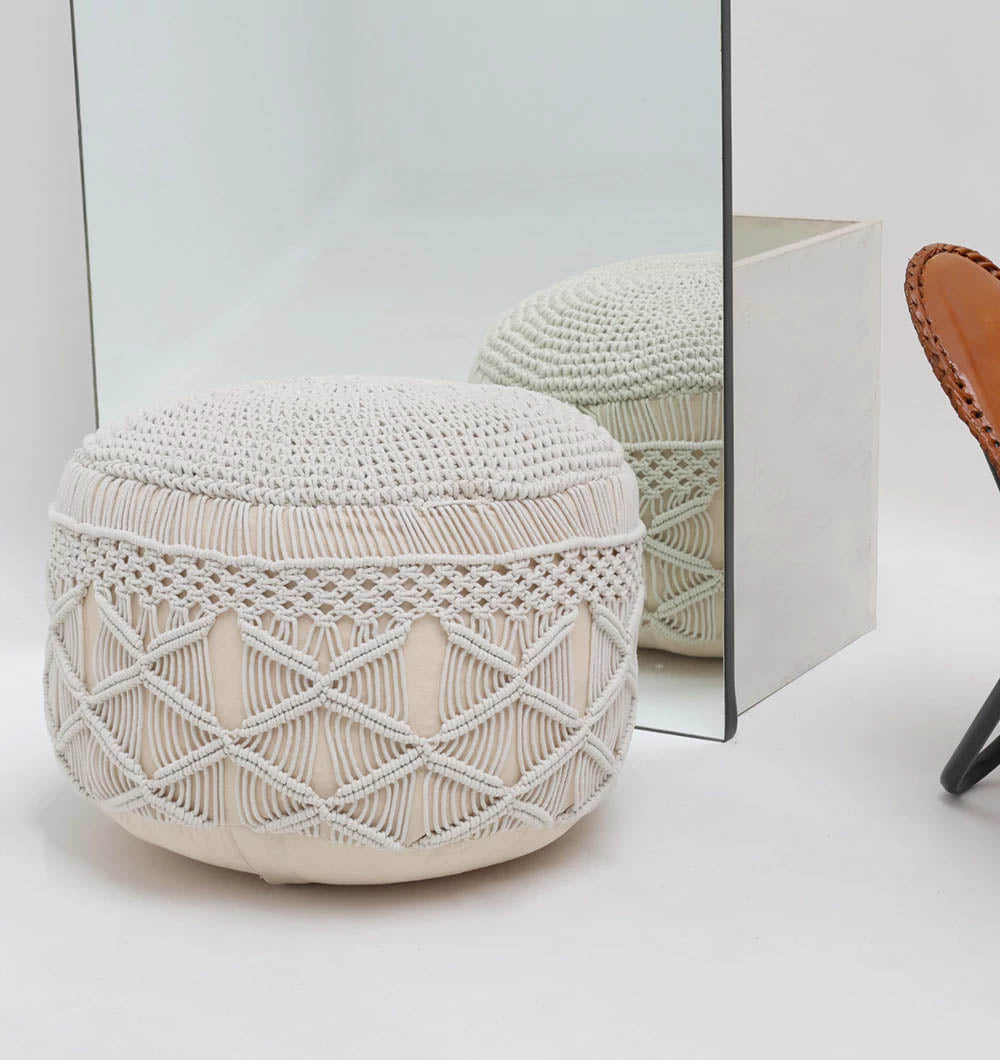 A RAMSHA HOME Boho Macrame Pouf White Round PF-157 with a geometric pattern sits on a white floor by a large mirror, which reflects the pouf. A portion of a brown leather chair is visible on the right side of the image.