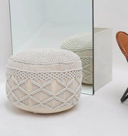 A RAMSHA HOME Boho Macrame Pouf White Round PF-157 with a geometric pattern sits on a white floor by a large mirror, which reflects the pouf. A portion of a brown leather chair is visible on the right side of the image.