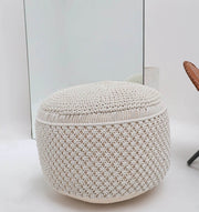 The RAMSHA HOME Boho Macrame Pouf White Round PF-158, featuring a knitted and woven texture, sits on a white floor before a large mirror, with part of a brown chair visible to the right.